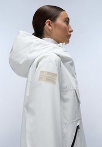 White waterproof jacket with a hood, side pockets, featuring a textured cream flag patch on the sleeve and smooth fabric.