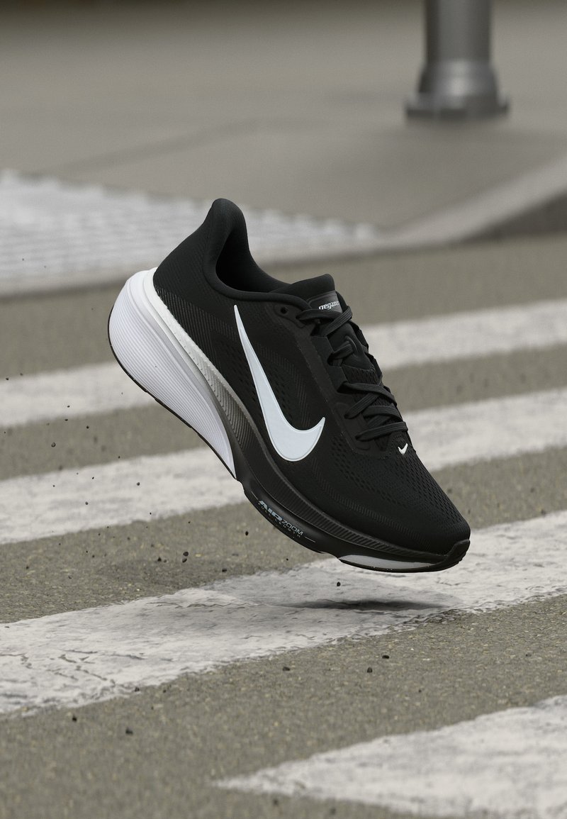 Μονόχρωμο μαύρο και λευκό παπούτσι Nike για τρέξιμο στον αέρα πάνω από διάβαση πεζών σε ασφάλτινο δρόμο.