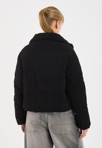 Svart kordfløyelskort jakke med bred krage og polstret tekstur. Har et rent bakdesign som komplimenterer grå denim bukser.