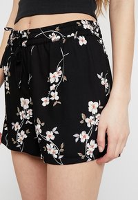 Svarta blommiga shorts i lättviktigt material. Har ett elastiskt midjeband och dragsko, prydda med vita blommor och gröna blad.