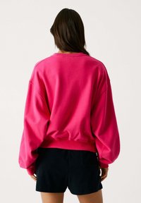 Sudadera rosa de gran tamaño con acabado texturizado, mangas anchas y dobladillo acanalado; combinada con pantalones cortos oscuros, capturando una silueta relajada.
