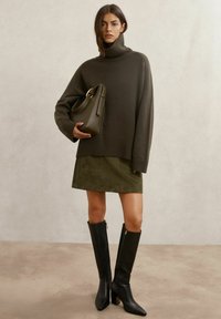 Pulover supradimensionat cu guler înalt, de culoare verde închis, fustă slim olive, cizme înalte din piele neagră cu fermoare aurii și o geantă de mână asortată.