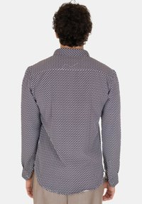 Uomo con capelli ricci che indossa una camicia a maniche lunghe con un motivo a catena nera e bianca, visto da dietro.