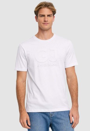 Uomo con capelli corti biondi che indossa una maglietta bianca a maniche corte con il logo "GJ GAUDI JEANS" in rilievo e jeans blu, in piedi davanti a uno sfondo grigio.