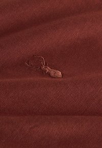 Karma sarkans audums ar mīkstu tekstūru, ar smalku izšūtu briedi kā logotipu. Virsma rāda smalku auduma pinumu.