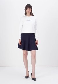 Felpa bianca a maniche lunghe con testo blu, abbinata a una gonna plissettata blu navy, indossata con tacchi alti neri su uno sfondo neutro.