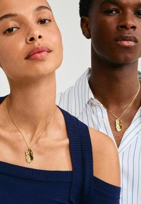 Colliers en ton d'or avec des pendentifs texturés en forme organique portés par des individus en tenue décontractée. L'arrière-plan est neutre.