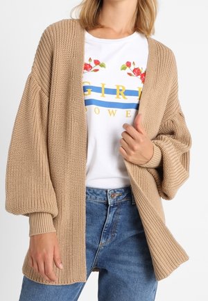 Vrouw die een beige grofgebreide cardigan draagt over een wit T-shirt met "Girl Power" en bloemmotief, gecombineerd met een blauwe spijkerbroek.