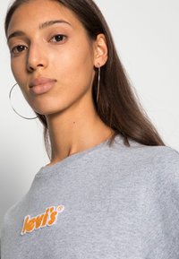 Szürke pamut póló, kerek nyakkivágással és narancssárga hímzett "Levi's" logóval. A modellen nagy karika fülbevaló van, és egyenes, hosszú haja van.