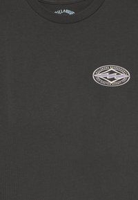 Camiseta de algodón negra con cuello redondo, que presenta un parche de logo circular en blanco y morado en el lado izquierdo del pecho. Textura suave, diseño casual.
