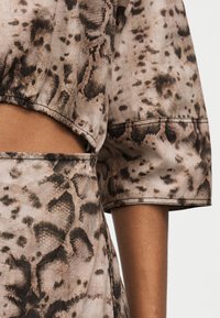 Κοντινή λήψη καφέ και μπεζ υφάσματος με σχέδιο φίδι, τοπ με cutout και μανίκι τριών τετάρτων, και ταιριαστά ψηλόμεσα κάτω κομμάτια στο μπράτσο και τη μέση ενός ατόμου.