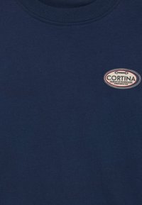 Tamno plavi majica kratkih rukava s okruglim izrezom, izrađena od mekog pamučnog materijala, sa kružnim izvezenim znakom označenim "CORTINA" koji sadrži crvene i bijele akcentne boje.