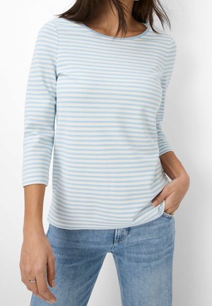 Vrouw draagt een lichtblauw-wit horizontal gestreept shirt met lange mouwen en een bootnek, gecombineerd met blauwe spijkerbroek, hand in de zak.