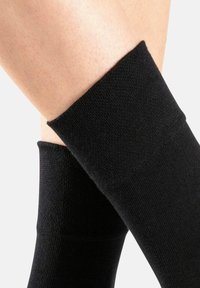 Schwarze knielange Socken aus weichem, dehnbarem Stoff. Glatte Textur mit einem gerippten Bunddesign. Formschlüssig und nahtlos am Knöchel.