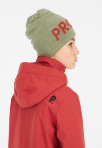 Giacca impermeabile rossa con cappuccio regolabile, caratterizzata da un logo nero sulla spalla sinistra. Abbinata a un berretto di lana verde con testo rosso.