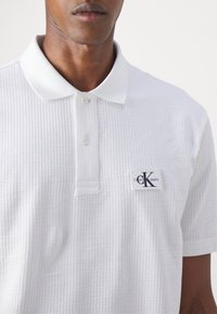 Homme portant un polo blanc texturé à manches courtes avec un patch logo Calvin Klein Jeans sur la poitrine.