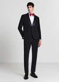 Schwarzer Smoking mit Satinrevers, kombiniert mit einem weißen Hemd und roter Fliege. Schlanke Hose und glänzende schwarze Anzugschuhe vervollständigen den Look.