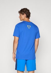 Plava pamučna majica s kratkim rukavima i okruglim izrezom. Ima žuti Nike logo na gornjem dijelu leđa. Uparena je s plavim šortsevima.