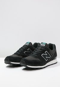 Un paio di sneaker nere New Balance 998 con suole bianche e marchio teal, esposte su una superficie bianca con uno sfondo grigio chiaro.