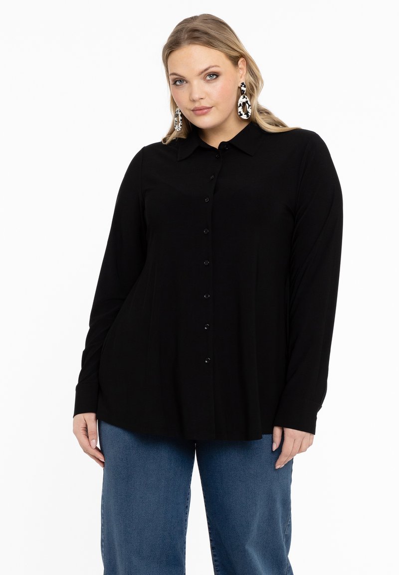 Femme aux longs cheveux blonds portant une chemise noire boutonnée, un jean bleu et des boucles d'oreilles pendantes à motifs noir et blanc sur un fond blanc.