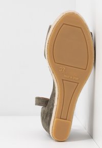 Semelle d’espadrille en daim gris avec semelle en caoutchouc beige, taille 37, fabriquée en Espagne, posée sur une surface blanche contre un mur blanc.
