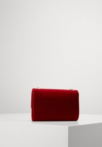 Borsetta clutch in velluto rosso con una texture liscia, design arrotolato e dettagli in metallo dorato. Posizionata su una superficie espositiva bianca.