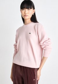 Světle růžová mikina vyrobená z jemné tkaniny, s kulatým výstřihem, žebrovanými manžetami a malým zeleným logem Lacoste na hrudi.