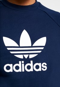 Námorně modrá mikina s kulatým výstřihem. Obsahuje velké bílé logo Adidas se třemi pruhy a ikonickým designem listu, které je výrazně zobrazeno.