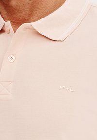 Jasnoróżowa koszulka polo z teksturowanej tkaniny, z klasycznym kołnierzykiem i trzema guzikami, z dyskretnym logo "P·L" na piersi.