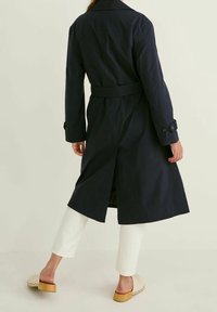 Navy trench coat with a belted waist, long sleeves, and buttoned cuffs. Features a vent at the back. Worn over white pants and light shoes.