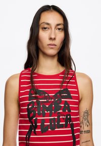 Czerwony top bez rękawów w paski o żebrowanej fakturze z dużym czarnym nadrukiem z napisem „BIMBA Y LOLA”. Posiada szerokie wycięcia na ramiona oraz okrągły dekolt.