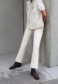 Top di cotone color crema con maniche a sbuffo corte e scollo a fiocco, abbinato a pantaloni a campana bianchi sporchi e stivaletti alla caviglia in suede marrone scuro.