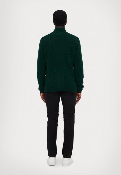 Green cable-knit sweater with high collar, black pants, and white sneakers. The fabric features a textured pattern and ribbed cuffs.
