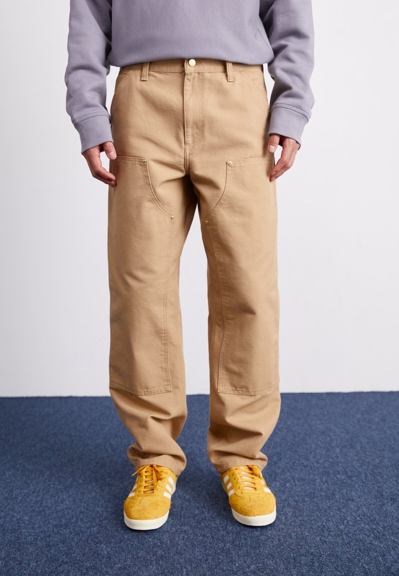 Pantalons cargo beiges avec poches avant, accessoires dorés et coupe décontractée, associés à des baskets jaunes avec des rayures blanches.
