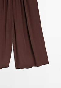 Pantalon marron à jambes larges avec une texture lisse, montré suspendu sur un fond blanc.