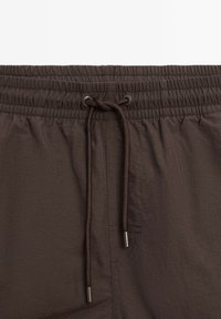 Bruine elastische tailleband met verstelbaar trekkoord. Glad, lichtgewicht stof met een subtiele glans en fijne stikdetails.