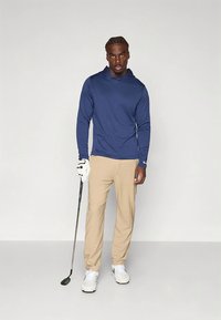 Mørkeblå hettegenser med lange ermer, beige bukser, hvite sko, holder en golfklubb; mykt stoff, avslappet passform, subtil merkevare på skjorten.