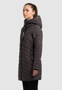 Young woman with brown hair in ponytail wearing a long, dark quilted winter coat with hood, standing in profile against a plain background.