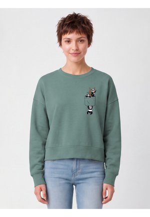 Young woman with short hair wearing a green sweatshirt with small cartoon pandas climbing on a chest pocket, paired with light blue jeans.