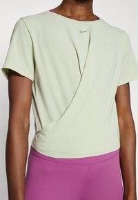 Kurzärmliges Oberteil aus Baumwollmischung in Hellgrün, mit einem verdrehten Vorderteil und einem dezenten Logo. Kombiniert mit hoch taillierten pinkfarbenen Leggings.