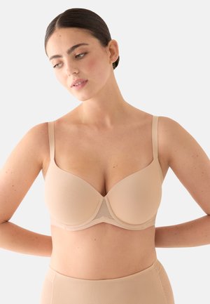 Mujer con cabello oscuro recogido, que lleva un sujetador beige sin costuras con aros y bragas de talle alto a juego, mirando hacia abajo y hacia un lado.