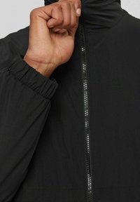Giacca nera con zip e colletto alto, polsini elasticizzati e una zip visibile con dettagli riflettenti sulla parte anteriore.