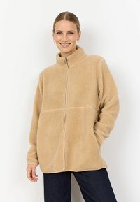 Beež fliisk jacket kõrge krae, täisfrontaalse lukk ja pikad, veidi mahukad varrukad, millel on tekstuuriline pind.