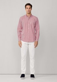 Chemise boutonnée à rayures rouges et blanches avec un col, des manches longues et une petite poche poitrine. Associée à un pantalon blanc et des chaussures foncées.