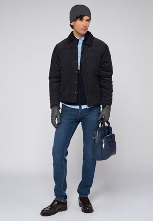 Giacca nera trapuntata, camicia blu, jeans blu scuro, guanti grigi e berretto abbinato. Mocassini marroni e una borsa blu completano l'insieme.