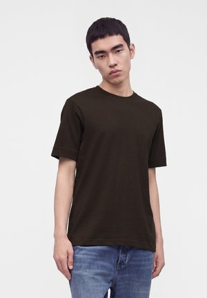 Joven con cabello corto y negro, vestido con una camiseta negra de manga corta lisa y vaqueros azules, de pie frente a un fondo claro y liso.