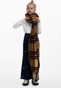 Ampia sciarpa scozzese multicolore in arancione, giallo e verde indossata sopra una maglia bianca a coste e una gonna di jeans blu con fiori ricamati.