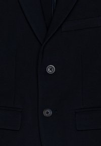 Námořnická modrá bunda s hladkým povrchem, klasickými klopami, dvěma kapsami a zapínáním na dva kulaté knoflíky.