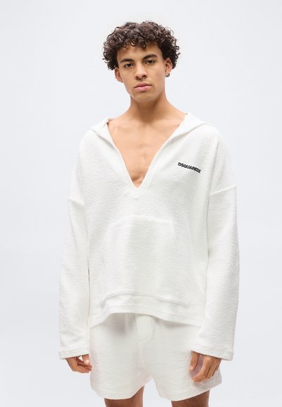 Ung man med lockigt hår som bär en överdimensionerad vit texturerad hoodie med djup V-ringning och matchande shorts, stående mot en enfärgad bakgrund.