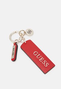 Llavero de cuero rojo que presenta una etiqueta rectangular grabada con "GUESS," una etiqueta secundaria con "EST. 1981," y herrajes metálicos.